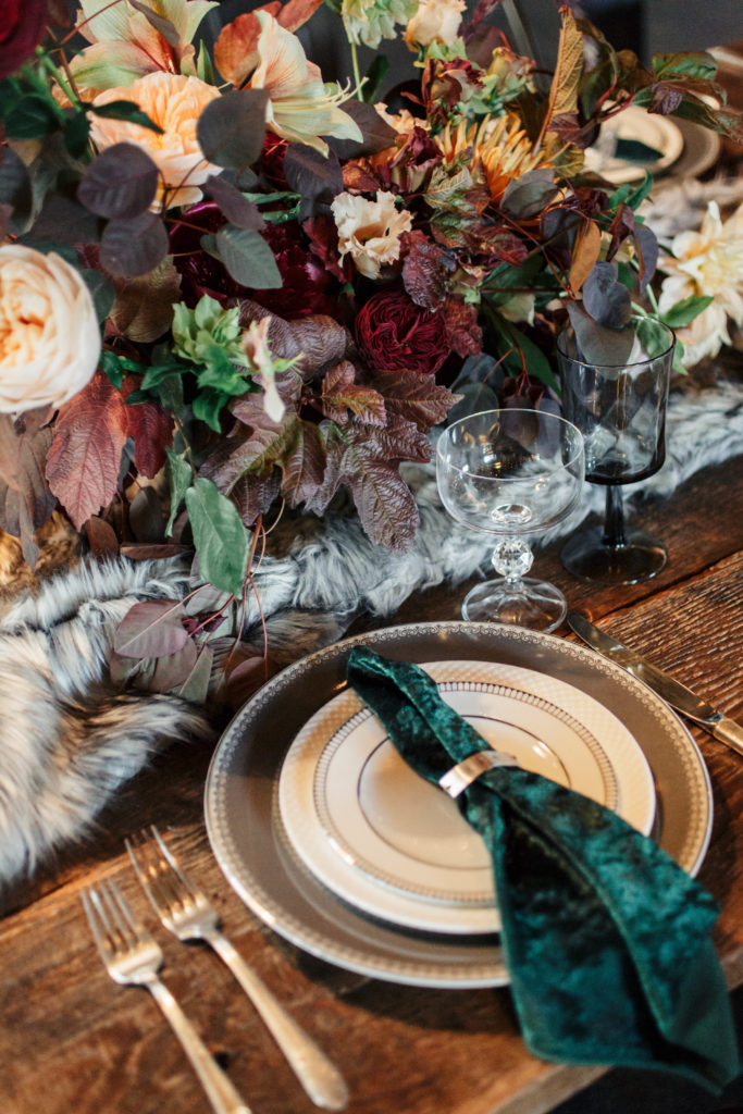 Holiday table decor on wooden table with velvet green napkin. Decorated with a faux fur table runner and burgundy and green florals