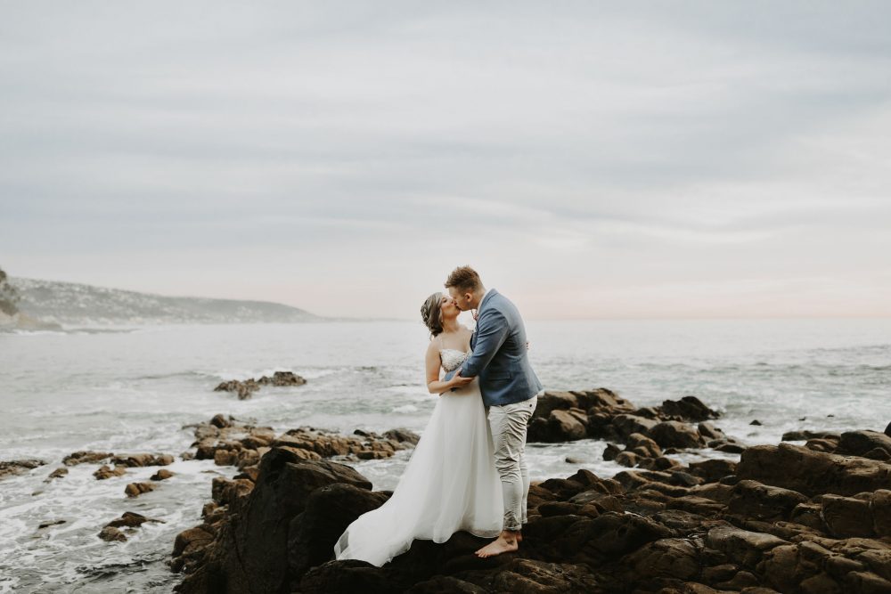 Bride and groom kiss on rocky cliff as waves crash behind them in Laguna Beach