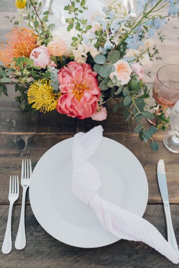 Long wooden table with white plates, knotted light pink napkins and silver flatware. Blush and clear wine glasses added a pop of color. The centerpieces were bright with pops of yellow, pink and white. Each table was decorated with a few airplants.