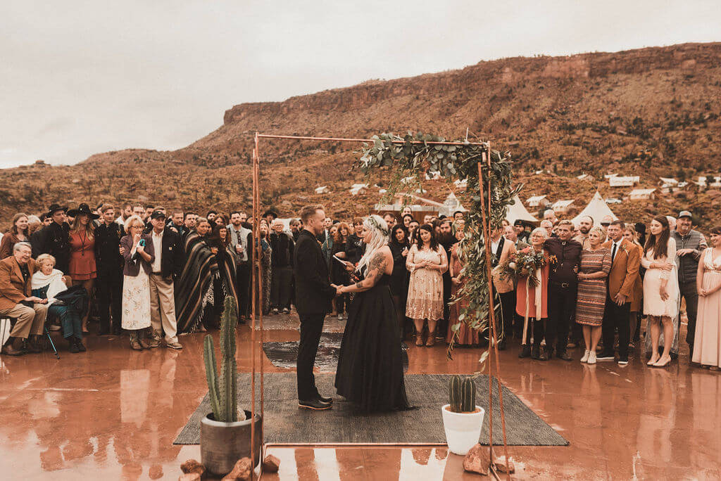 Bride and groom in all black in front of copper ceremony arch with greenery. Ceremony and reception at Zion Under Canvas for a glamping wedding