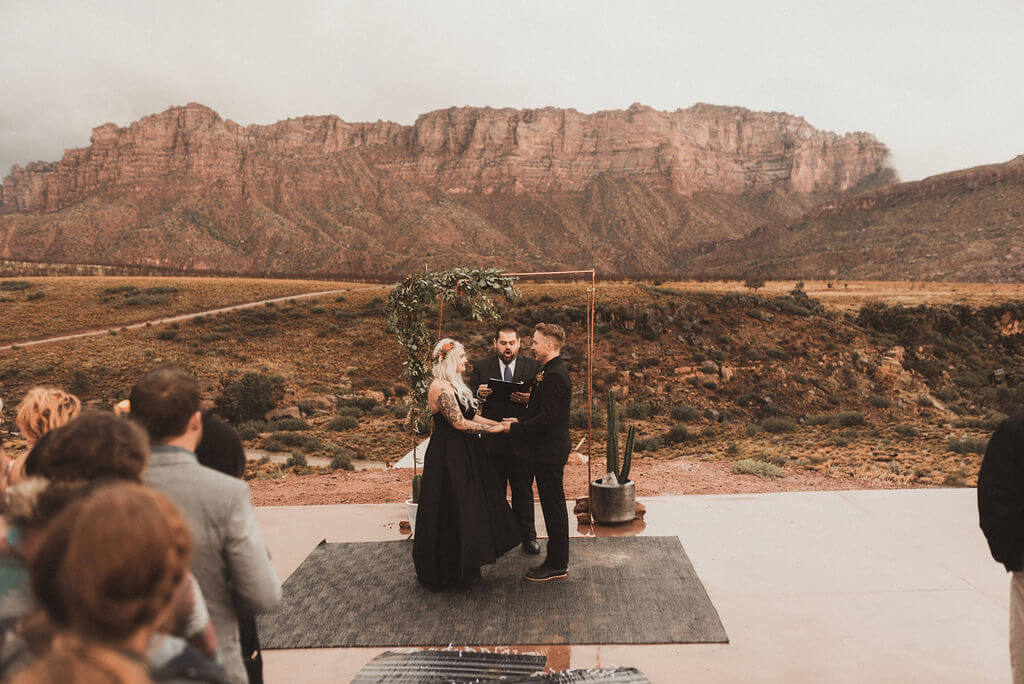 Bride and groom in all black in front of copper ceremony arch with greenery. Ceremony and reception at Zion Under Canvas for a glamping wedding