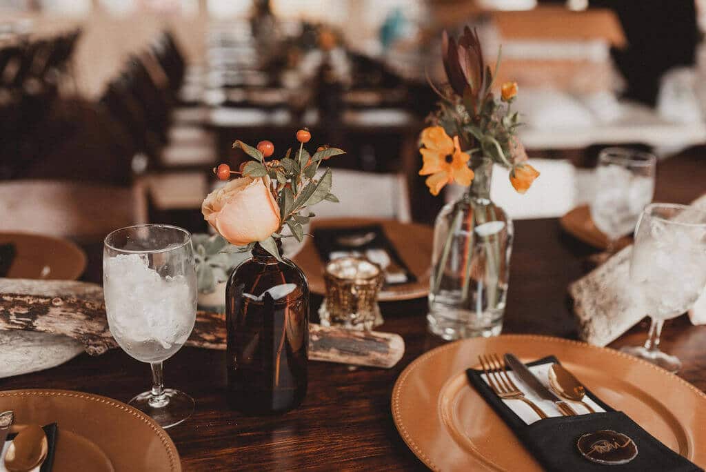 Wedding reception table with gold charger, black napkin, amber bud vases and dark flowers. Driftwood pieces on long wooden tables, pampass grass, king protea and cacti.