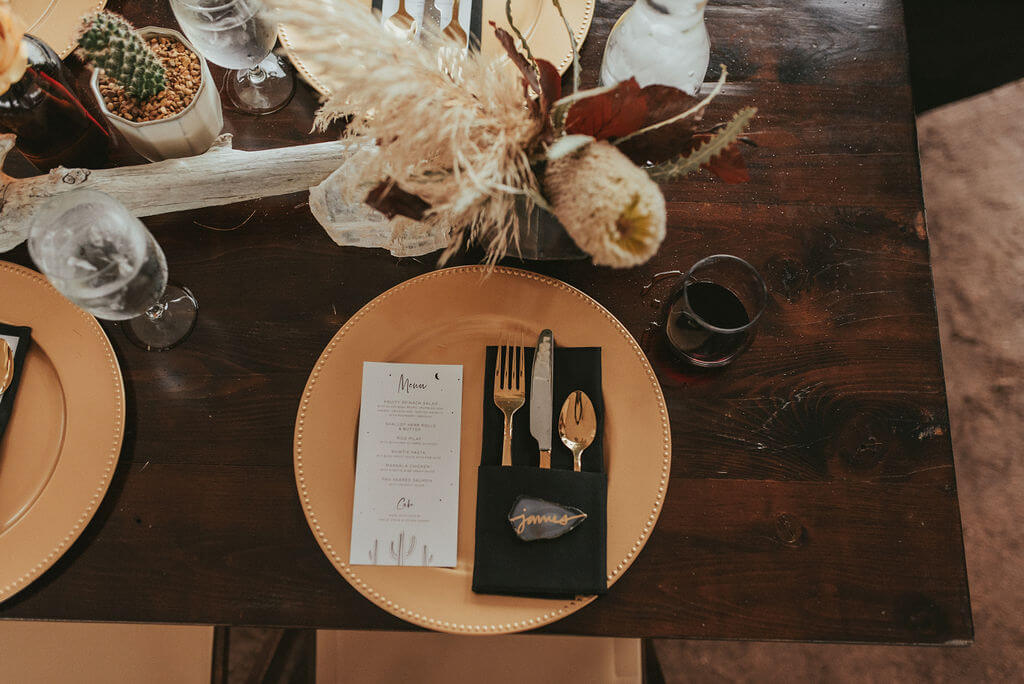 Wedding reception table with gold charger, black napkin, amber bud vases and dark flowers. Driftwood pieces on long wooden tables, pampass grass, king protea and cacti.