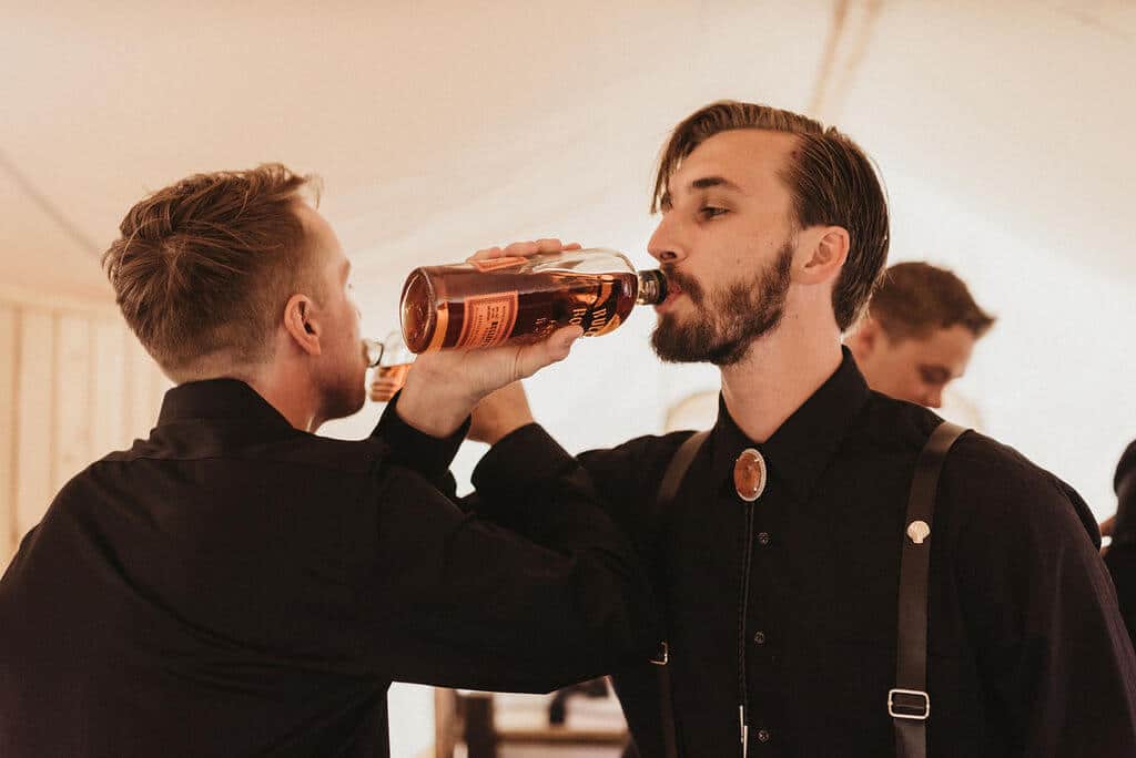 Groom and groomsmen sharing a bottle of whiskey before the wedding. Groom wearing an all black suit and shirt.